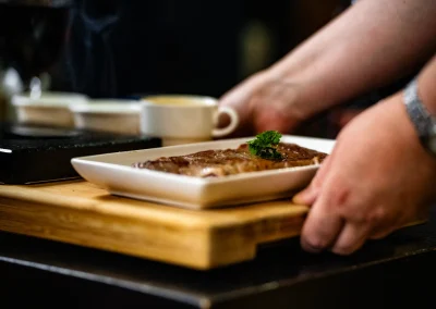 A server presenting a cooked steak with parsley garnish on a wooden board at the table.